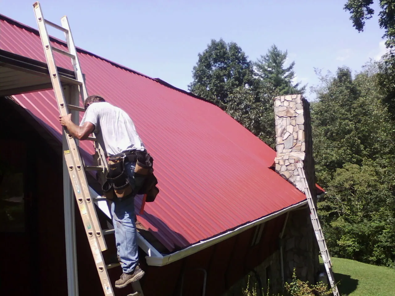 Roofer on a ladder beside a red metal roof and stone chimney — Soffit & Fascia Repair by Roofing Brothers in Rancho Santa Fe